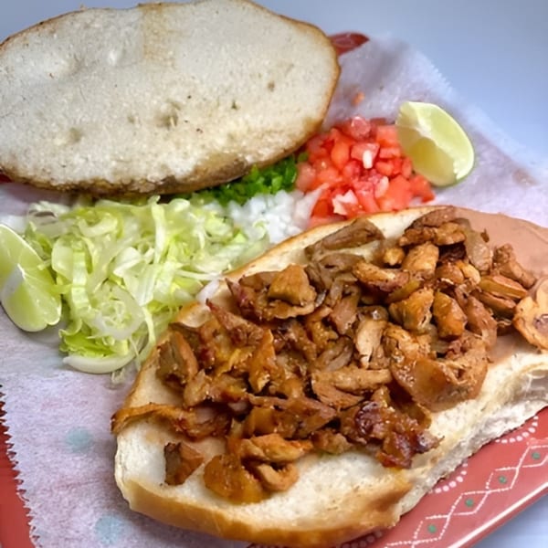 Open-faced torta with seasoned meat, shredded lettuce, diced tomatoes, onions, and lime wedges.